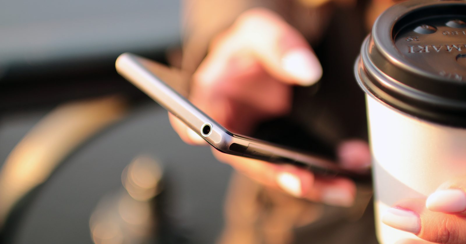 Osoba trzymająca smartfon nad stołem, obok filiżanka kawy i notatnik.