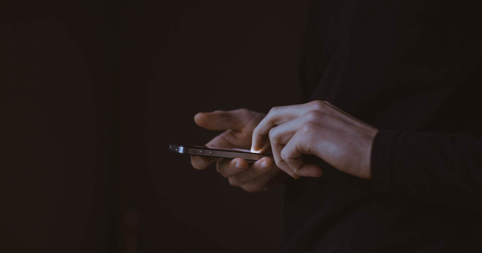 Dłonie trzymające smartfon w ciemności, ekran podświetla palce użytkownika.